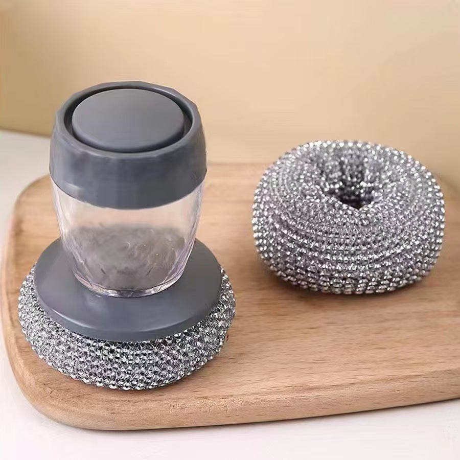 Soap Dispensing Steel Brush with gray lid and base and metallic scrubber on a wooden board.