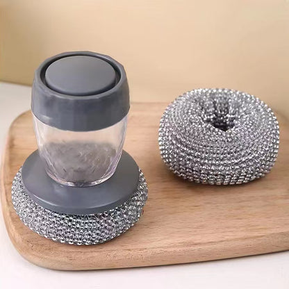 Soap Dispensing Steel Brush with gray lid and base and metallic scrubber on a wooden board.